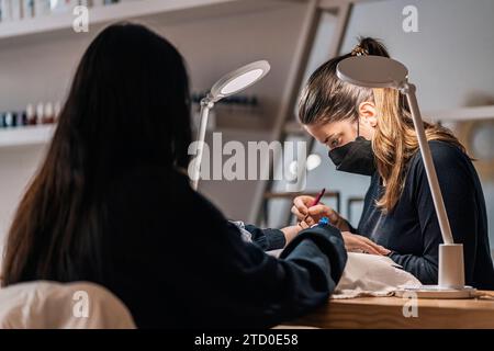 Nicht erkennbare Kundin, die sich einer Nagelbehandlung durch Kosmetikerin mit Maske im Manikürstudio unterzieht Stockfoto