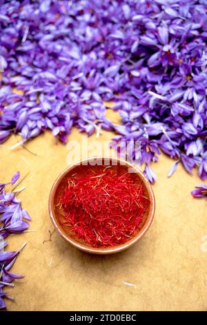 Von oben ist eine Tonschale gefüllt mit zarten roten Safranfäden auf einem Holztisch inmitten violetter Blütenblätter, die den Kontrast und das Endprodukt des h hervorhebt Stockfoto