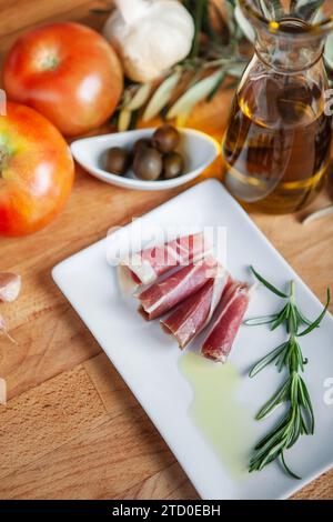 Ein Holztisch bietet mediterrane Zutaten mit Schwerpunkt auf iberischem Schinken, frischen Tomaten, Knoblauch, Oliven und einer Flasche Olivenöl mit Stockfoto