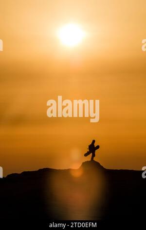 Ganzkörper der Silhouette nicht erkennbare Person mit Snowboard, die auf dem Berg vor dem Himmel steht und während des Sonnenuntergangs in Japan im Winter helle Sonne hat Stockfoto