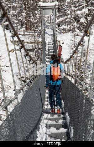 Ganzkörper-Rückansicht eines anonymen Mannes in warmer Kleidung und Skiausrüstung, der im Winter auf einer Hängebrücke in Zermatt, Schweiz, läuft Stockfoto