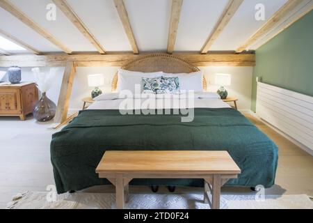 Ein einladendes Schlafzimmer im Dachgeschoss mit freiliegenden Holzbalken, einer üppigen grünen Federdecke und rustikalen Möbeln, die eine warme Atmosphäre schaffen. Stockfoto