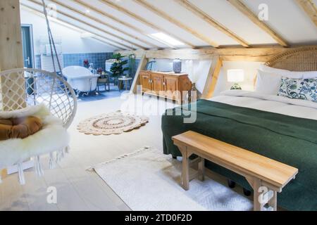 Geräumiges Schlafzimmer im Dachgeschoss mit Holzbalkendecke, gemütlichem Bett und offenem Bad mit freistehender Badewanne. Stockfoto