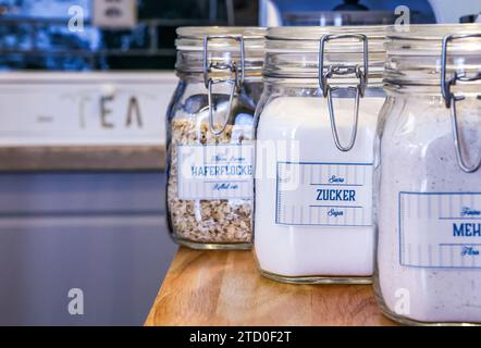 Drei Vorratsdosen aus Glas mit Metallverschlüssen und Etiketten für Hafer, Zucker und Mehl, ordentlich auf einem hölzernen Küchenregal angeordnet. Stockfoto