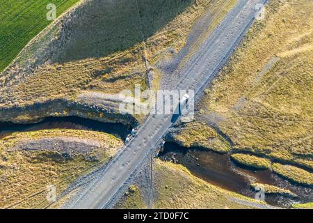 Ein Luftbild, das ein Auto auf den Serpentinenpfaden einer kurvenreichen Straße durch die grüne und braune Landschaft des isländischen Hochlands einfängt Stockfoto