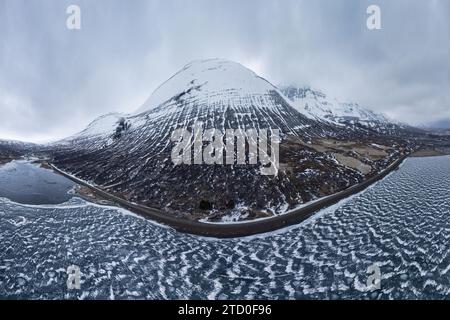Panoramablick auf einen majestätischen, leicht mit Schnee bedeckten Berg neben einem gefrorenen See in der isländischen Wildnis während des Winters. Stockfoto