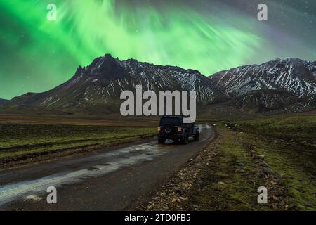 Eine atemberaubende Aurora Borealis erleuchtet den Nachthimmel über einer zerklüfteten Bergkette, während ein einsames Fahrzeug auf einer kurvenreichen Straße in Island fährt Stockfoto