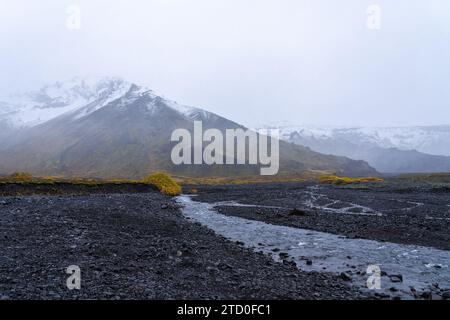 Ein ruhiger Bach schlängelt sich durch eine schwarze Kiesellandschaft mit nebelumhüllten Bergen im Hintergrund in Island. Stockfoto