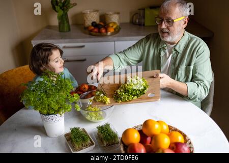 Blick aus der Höhe auf den älteren Großvater, der eine Brille trägt und Salat in eine Schüssel legt, während er mit der Enkelin zu Hause Salat zubereitet Stockfoto