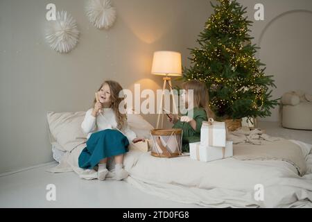 Zwei junge Mädchen, die auf einem gemütlichen Bett neben einem wunderschön dekorierten Weihnachtsbaum mit leuchtenden Lichtern Urlaub machen. Stockfoto