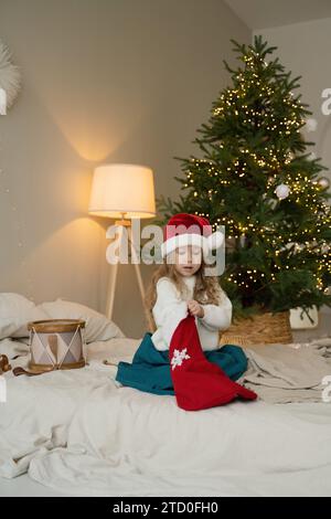 Ein junges Mädchen in einem Weihnachtsmann-Hut sitzt auf einem Bett und spielt sanft mit ihrem Hut, mit einem beleuchteten Weihnachtsbaum im Hintergrund. Stockfoto