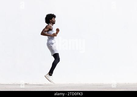 Seitenansicht eines entschlossenen jungen aktiven afroamerikanischen Sportlers in Sportbekleidung, der während des Fitnesstrainings auf der Straße gegen die weiße Wand springt Stockfoto