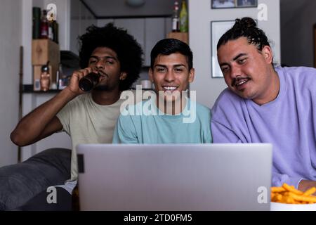 Fröhliche junge männliche Freunde in lässigen Videoanrufe über kabellosen Computer im Wohnzimmer während der Freizeit Stockfoto