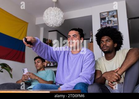Multirassische ernsthafte männliche Freunde in legeren Kleidern, die das Smartphone benutzen und zusammen Fernsehen, während sie im Wohnzimmer sitzen, mit kolumbianischer Flagge im Rücken Stockfoto