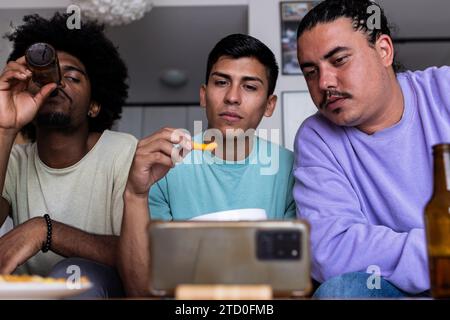 Niedriger Blickwinkel auf ernsthafte junge männliche Freunde, die am Wochenende Bier und Snacks trinken, während sie Sportereignisse über das Mobiltelefon im Wohnzimmer beobachten Stockfoto