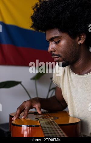 Nahaufnahme eines fokussierten jungen Mannes mit Afro-Haaren, der Akustikgitarre spielt, während er am Wochenende zu Hause gegen kolumbianische Flagge sitzt Stockfoto