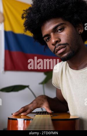 Nahaufnahme eines fokussierten jungen Mannes mit Afro-Haaren, der Akustikgitarre spielt, während er am Wochenende zu Hause gegen kolumbianische Flagge sitzt Stockfoto