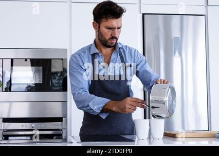 Konzentrierter bärtiger, erwachsener Küchenchef in Schürze, der in der Küche nach unten blickt und während der Frühstücksvorbereitung Zutaten aus dem Topf in Glas gießt Stockfoto