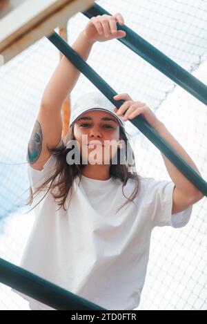 Eine junge Frau mit einem Tattoo auf dem Arm posiert lässig hinter einem grünen Metallzaun und sieht entspannt und selbstbewusst in einem einfachen weißen T-Shirt aus Stockfoto
