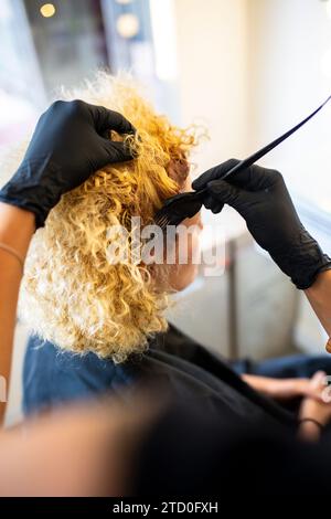 Ein Friseur trägt schwarze Handschuhe und trägt in einem professionellen Salon sorgfältig Haarfarbe auf das lebhafte lockige blonde Haar eines Kunden auf. Stockfoto