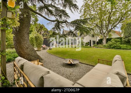 Komfortables Sofa mit Feuerstelle auf Terrasse mit grasbewachsenem Rasen umgeben von üppigen Bäumen mit moderner Villa im Hintergrund Stockfoto
