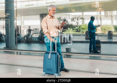 Der ganze Körper eines bärtigen älteren Mannes in Brillen und Gelegenheiten, der Nachrichten über das Smartphone überprüft, während er am Fenster des modernen Flugplatzes steht Stockfoto