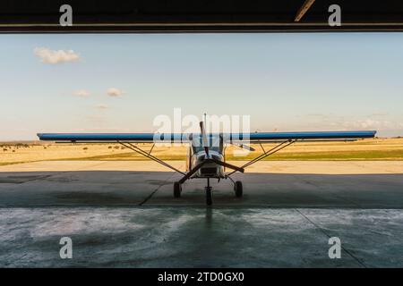 Ein einmotoriges Flugzeug wird im Hangar in Ruhe gefangen genommen, wobei die offene Tür einen klaren Himmel und die Start- und Landebahn dahinter einrahmt Stockfoto
