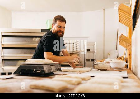 Fokussierter bärtiger, kaukasischer männlicher Bäcker, der in Casuals gekleidet ist und rohen Teig auf dem Tisch für die Herstellung von Gebäck in der Küche im Backhaus kneten Stockfoto