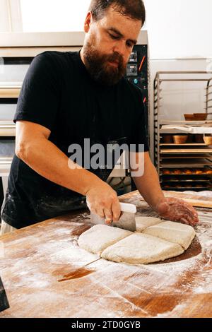 Fokussierter seriöser, reifer männlicher Koch in der Freizeit, der frischen Teig mit einem Cutter auf Holztisch für die Zubereitung von Gebäck in der Küche im Backhaus schneidet Stockfoto