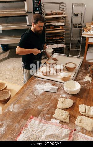 Hochwinkelansicht eines fokussierten männlichen Bäckers, der Mehl auf ungekochtem Teig in Schalen am Holztisch siebt, während er in der Küche in der Bäckerei frisches Gebäck zubereitet Stockfoto