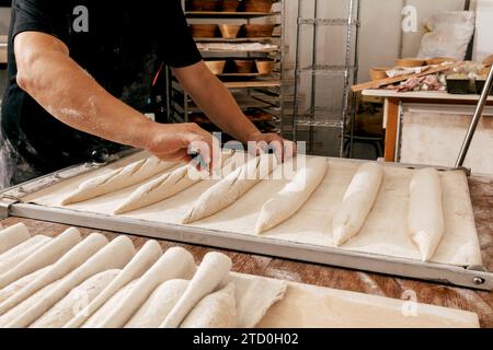 Beschneiden Hände eines nicht erkennbaren männlichen Küchenchefs, der in Casuals gekleidet ist und ungekochte Brotlaibe mit einem Messer im Tablett vor dem Backen am Tisch in der Küche einschneidet Stockfoto