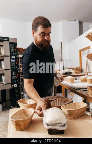 Bärtiger ausgereifter männlicher professioneller Bäcker, der ungekochtes Sauerteigbrot in Formen mit Kunststoff auf Holztisch in der Küche im Backhaus bedeckt Stockfoto