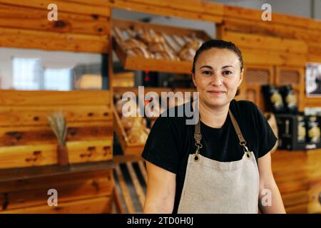 Porträt einer selbstbewussten positiven Verkäuferin in Schürze mit Händen auf der Theke, die neben frisch gebackenem Brot auf Holzregalen in Bak steht Stockfoto