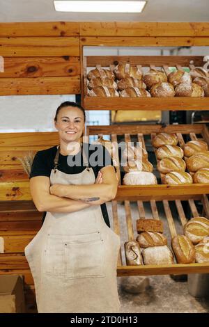 Porträt einer selbstbewussten, positiven Verkäuferin in Schürze mit überkreuzten Armen, die von frisch gebackenem Brot auf Holzregalen in der Bäckerei stehen Stockfoto