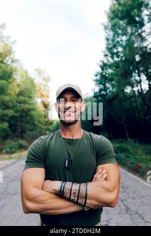 Porträt eines selbstbewussten, reiferen männlichen Kommandos mit überkreuzten Armen und Tätowierung in der Kappe lächelnd in die Kamera, während er auf der Straße gegen den Himmel im Wald steht Stockfoto