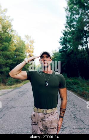 Ein selbstbewusster, reifer Militärsoldat trägt eine Tarnhose und ein grünes T-Shirt, der vor Stolz grüßt, während er auf der Straße gegen den Himmel im Wald steht Stockfoto