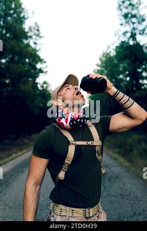 Durstiger Marine-Kommandant mittleren Alters trägt ein grünes T-Shirt und eine Kappe, die Wasser aus der Flasche trinkt, während er auf der Straße im Wald steht Stockfoto