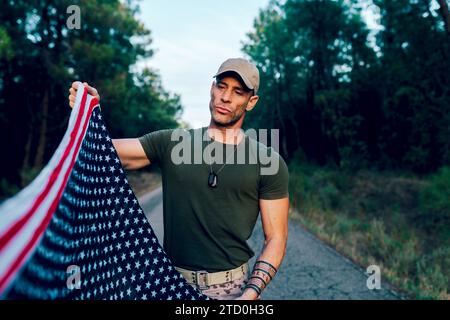 Sondereinsatzkommando mittleren Alters in Uniform und mit Blick auf die amerikanische Flagge, während er auf der Straße gegen Bäume im Wald steht Stockfoto