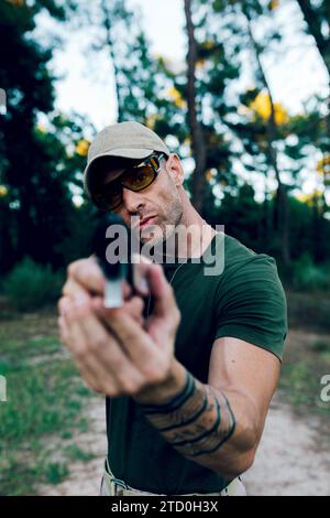 Armee-Kommando mittleren Alters in Kappe und Brille, die in die Kamera blickt, während sie mit der Waffe im Wald gegen Sonnenuntergang zielt Stockfoto