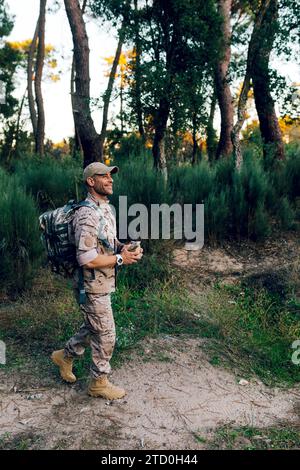 Seitenansicht eines glücklichen, Reifen Militärsoldaten in Tarnuniform mit Kappe und Rucksack, der bei Sonnenuntergang im Wald steht Stockfoto