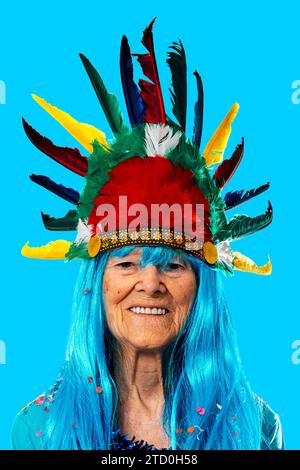 Porträt der lächelnden Seniorin blau in Perücke mit Konfetti mit indischem Kopfschmuck mit bunten Federn, während sie vor blauem Hintergrund und l steht Stockfoto