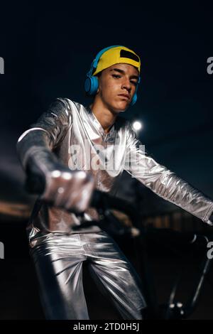 Niedriger Winkel eines jungen Mannes in silbernem Kostüm mit Handschuhen und gelber Kappe, der in die Kamera blickt, während er mit Kopfhörern bis zum Ohr und an den Holm fährt Stockfoto
