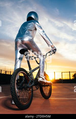 Rückansicht eines anonymen Mannes in Helm und silbernem Kostüm, der wegschaut, während er auf leerem Boden Fahrrad fährt und Tricks gegen bewölkten Sonnenuntergang macht b Stockfoto
