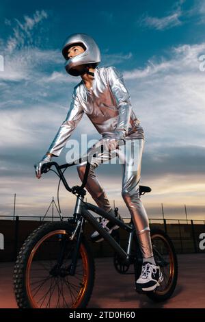 Niedriger Winkel Ganzkörper Seitenansicht eines Mannes mit Helm und silbernem Kostüm, der wegschaut, während er auf leerem Boden Fahrrad fährt und Tricks gegen Wolken macht Stockfoto