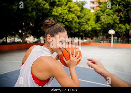 Lächelnde, fitte junge Frau in Sportbekleidung, die Basketball hält, während sie mit einem männlichen Freund auf dem Spielplatz mit Bäumen im Hintergrund spielt Stockfoto
