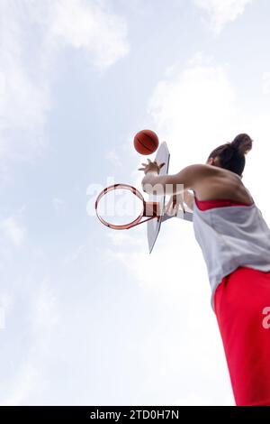 Von unten aus springt eine nicht erkennbare Basketballspielerin in Aktivkleidung und wirft den Ball in den Korb vor bewölktem Himmel Stockfoto