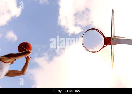 Von unten aus springt eine nicht erkennbare Basketballspielerin in Aktivkleidung und wirft den Ball in den Korb vor bewölktem Himmel Stockfoto