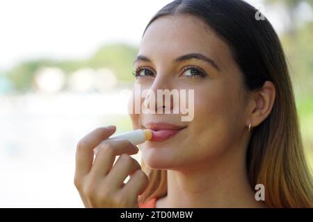 Nahaufnahme einer lächelnden jungen Frau, die draußen Sonnenschutz auf ihrer Lippe aufträgt Stockfoto