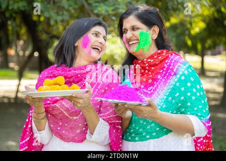 Zwei glückliche Indianerinnen tragen eine weiße Kurta mit einem Farbpuder oder gulal und Laddu, die das holi-Festival im Park oder Garten feiern. Stockfoto