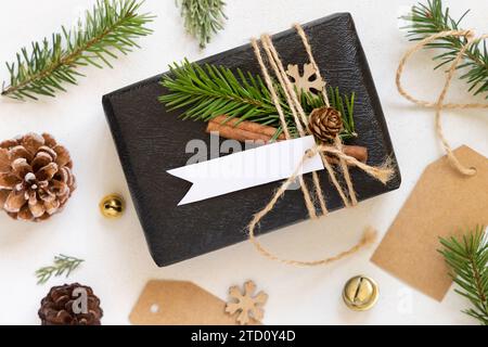 Schwarzes Geschenk mit weihnachtlichem horizontalen Geschenkanhänger, Tannenzweigen, Tannenzapfen und Weihnachtsdekoration, Draufsicht. Winterkomposition mit leerem Etikett Stockfoto